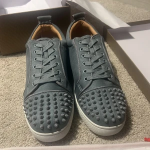 Grå Christian Louboutin sneakers med nitar - Snygga grå sneakers från Christian Louboutin med coola nitar på tån och klassisk röd sula. Skorna har snörning, rund tå och är tillverkade i mocka med vita kanter och detaljer. Perfekta för dig som vill sticka ut med exklusiv streetstyle.