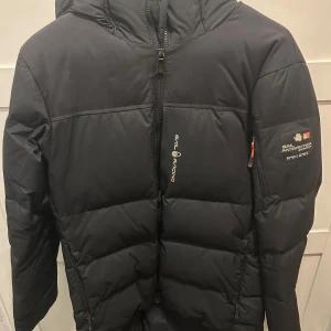 Svart dunjacka Sail Racing - Svart dunjacka från Sail Racing med huva och dragkedja framtill. Jackan har logga på bröstet och ett Sail Antarctica-märke på ärmen. Puffig modell som håller dig varm under kalla dagar. Perfekt för dig som vill ha en stilren och funktionell vinterjacka.