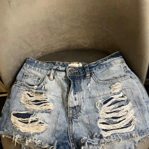Blå slitna jeansshorts med knappar - Riktigt snygga blå jeansshorts från Perfect Jeans med hög midja och flera stora slitningar framtill och baktill. Shortsens råa kanter och fransar ger en cool vibe. Klassisk femficksmodell och knäppning med knappar framtill. Perfekta för sommaren!