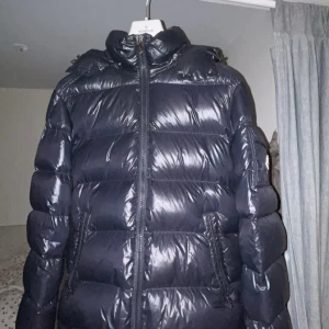 Marinblå Moncler dunjacka med huva - Säljer en marinblå, glansig dunjacka från Moncler med huva och dragkedja framtill. Jackan har Moncler-logga på ärmen, två fickor med dragkedja och klassisk pufferstil. Perfekt för kalla dagar när du vill hålla stilen och värmen. Storlek 4 Xl passar även L