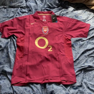 Arsenal Highbury Henry 14 Nike tröja L - Säljer en ikonisk Arsenal Highbury 1913-2006 matchtröja i vinröd med guldfärgade detaljer och tryck. Tröjan har korta ärmar, klassisk krage, broderat klubbmärke och Henry 14 på ryggen. Tröjan är i helt nyskick och har även kvar lappar .Storleken är L och passar för någon som är 175-180 cm lång. Vid minsta fråga eller om du vill ha mer bilder på tröjan hör av dig.