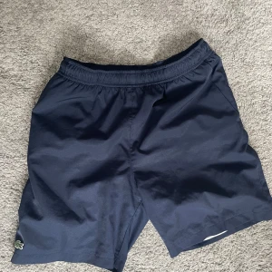 Mörkblå shorts från Lacoste - Mörkblå shorts från Lacoste med klassisk krokodillogga på benet. De har elastisk midja och är tillverkade i ett lätt, syntetiskt material som passar perfekt för varma dagar. Enkel och sportig design med normal passform.