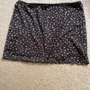 Svart blommig kortkjol med spetskant - Supersöt svart kortkjol med små blommor i ljusrosa och vit. Kjolen har en fin spetskant nertill och en tunn, luftig känsla. Perfekt som topp också! Aldrig använd