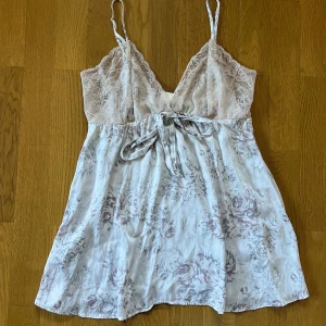 Blommig spetstopp från JJ's Benson - Supersöt babydoll topp med tunna axelband, spets upptill och blommigt mönster i ljusa toner. Toppen har v-ringning och ett knytband under bysten. Materialet är mjukt och svalt, perfekt för varma dagar.