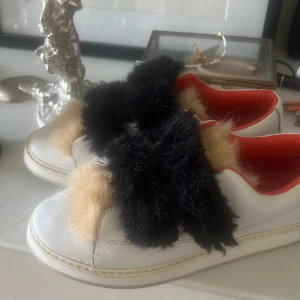 Vita sneakers med pälsdetaljer - Unika vita sneakers med fluffiga beige och svarta pälsdetaljer över tån och snörningen. Insidan är röd för en cool kontrast. Skorna har rund tå och platt sula, perfekta för dig som vill sticka ut med din stil. Pris kan diskuteras!