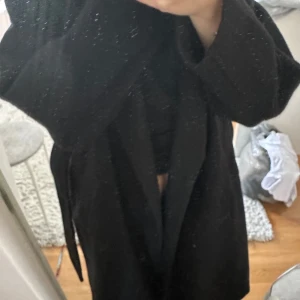 Svart kappa med bälte i ullmix - Säljer en svart kappa i ullmix med bälte i midjan och stora fickor. Kappan har en klassisk, rak passform och långa ärmar. Perfekt för dig som vill ha en stilren och tidlös look under höst och vinter.