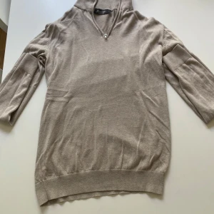 Beige polotröja Dressmann Supima S - Beige polotröja från Dressmann i mjuk Supima-bomull. Tröjan har trekvartsärm, ribbade muddar och en klassisk polokrage med liten dragkedja framtill. Perfekt för dig som gillar stilren och enkel look.