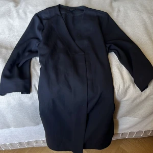 Svart minimalistisk klänning från COS - Säljer en svart, stilren klänning från COS i storlek 42. Jackan har en rak och minimalistisk design med omlottstängning och ärmar. Perfekt för dig som gillar en clean och modern look. Materialet känns mjukt och har en snygg struktur.