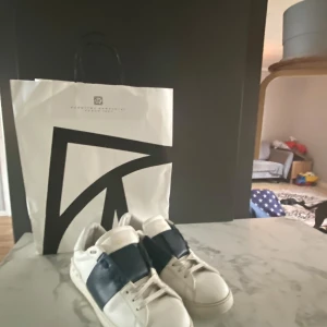 Valentino Open sneakers vit/marinblå - Snygga Valentino Open sneakers i vitt skinn med bred marinblå läderdetalj över vristen. Klassisk låg modell med rund tå, platt sula och diskret logga på hälen. Perfekta för dig som gillar stilrena och exklusiva sneakers med en twist.