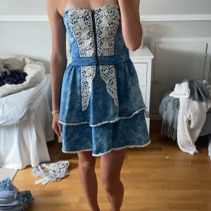 Blå jeansklänning med spetsdetaljer - Supersöt kort jeansklänning i ljusblå tvätt med vita spetsdetaljer framtill och längs volangkanten. Axelbandslös modell med dragkedja framtill och smock i ryggen. Den har tyvärr ingen lapp så vet inte märke eller storlek men den passar mig som vanligtvis har storlek S. Nästan aldrig använd så i jättebra skick! 