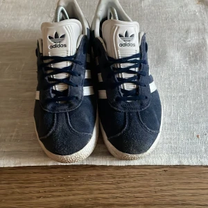 Adidas Gazelle mörkblå sneakers - Snygga Adidas Gazelle sneakers i mörkblå mocka med vita detaljer och klassiska tre ränder på sidan. Skorna har vit plös och hälkappa med Adidas-logga, samt snörning framtill. Perfekta för dig som gillar retrostil och streetwear.stora i storleken!!!! 