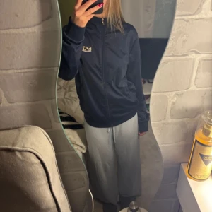 Mörkblå vindjacka från EA7 Emporio Armani - Snygg mörkblå vindjacka från EA7 Emporio Armani med dragkedja framtill och hög krage. Jackan har logga på bröstet och diskreta ränder på ryggen. Tillverkad i polyester, perfekt för dig som gillar sportig och clean stil.