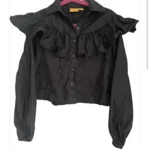 Ny svart volangblus från Gina Tricot - Ny svart blus med volanger över axlar och bröst, från Gina Tricot. Modellen har långa ärmar, en liten krage och knappar framtill. Perfekt för dig som vill ha något fint och svalt nu till sommaren. Finns i Kållered. Betalas med Swish. 