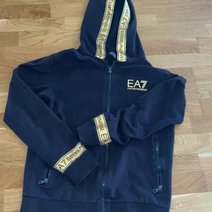 Svart EA7 hoodie med guldiga detaljer - Svart hoodie från EA7 Emporio Armani med guldiga logoband på huva och ärmslut. Tröjan har dragkedja framtill, två fickor med dragkedja och huva. Snygg streetstil och perfekt för dig som gillar exklusiva märken.