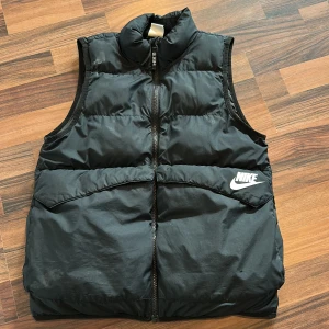 Svart dunväst från Nike - Svart dunväst från Nike med hög krage och dragkedja framtill. Västen har ett stilrent, quiltat mönster och en vit Nike-logga på bröstet. Perfekt för lager-på-lager och håller dig varm under kyliga dagar.