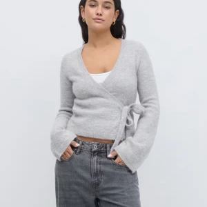 Grå omlottkofta från Gina Tricot - Supermjuk grå omlottkofta från Gina Tricot i ullblandning. Koftan har långa ärmar, v-ringning och knyt i sidan för en snygg passform. Perfekt att slänga över ett linne eller t-shirt för en chill look.