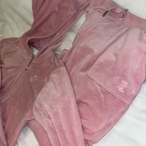 Rosa velour hoodie från Juicy Couture - Supermjuk rosa hoodie från Juicy Couture i klassisk velour med dragkedja och huva. Ikonisk JC-brodyr på bröstet och snygga detaljer vid dragkedjan. Perfekt för en chill och trendig look.