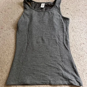 Mörkgrå linne - Snygg mörkgrå linnetopp från Lindex med klassisk rund halsringning. Toppen är ärmlös och har en enkel, stilren design som passar perfekt till jeans eller kjol. Mjuk och skön bomullsblandning som känns bekväm hela dagen. Har lite spets på ryggen, aldrig använd 