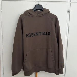 Brun Essentials hoodie med huva - Säljer en brun oversized hoodie från Essentials med stor svart logga framtill. Tröjan har huva, känguruficka och mjukt material i bomullsblandning. Perfekt för en chill och avslappnad stil. Essentials-märke även på huvan.