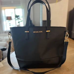 Svart axelväska från Michael Kors - Stilren svart axelväska från Michael Kors i skinn med guldfärgade detaljer och logga framtill. Väskan har både handtag och avtagbar axelrem, perfekt för att bära på olika sätt. Klassisk design som passar till många outfits.