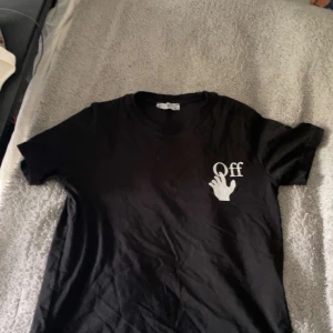 Svart Off-White t-shirt med tryck - Svart t-shirt från Off-White med vit handgrafik och texten 'Off' på bröstet. Baksidan har det ikoniska Off-White-korset med fototryck i vitt. Klassisk passform och rund hals, perfekt för dig som gillar streetwear och snygga detaljer.