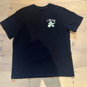 Svart Stüssy t-shirt med blomtryck - Svart t-shirt från Stüssy i storlek XL med coolt blomtryck och Stüssy-logga både fram och bak. T-shirten har korta ärmar och är gjord i mjuk bomull. Perfekt för dig som gillar streetwear och vill sticka ut med snygga detaljer.