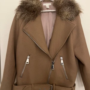 Beige kappa med fuskpäls från H&M storlek 36 - Säljer en beige kappa från H&M med bred krage i fuskpäls, snedställd dragkedja och två fickor med dragkedja. Kappan har bälte i midjan och är perfekt för kyligare dagar.   Lite nopprig men jättefin. Använd 2-3 gånger.  Står Storlek 36 men kan även användas av 38.  Pälsen är ej avtagbar!