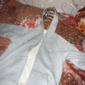 Grå Burberry hoodie med klassiskt foder - Snygg grå hoodie från Burberry med dragkedja och luva . Insidan av huvan har det  rutiga Burberry-mönstret i beige, svart och rött. Tröjan har snörning vid huvan och två fickor framtill. Perfekt för en chill och stilren look.
