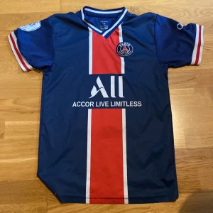 PSG Mbappé matchtröja blå röd 176 - Säljer en officiell PSG fotbollströja med Mbappé och nummer 7 på ryggen. Tröjan är blå med röd och vit rand mitt på, kortärmad och har klubbmärke på bröstet. Tillverkad i 100% mikrofiber, perfekt för dig som älskar fotboll och Paris Saint-Germain.