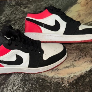 Nike Air Jordan 1 Low svart/vit/röd - Snygga Nike Air Jordan 1 Low sneakers i svart, vitt och rött. Klassisk design med svart swoosh, perforerad tå och lågt skaft. Ovandel i skinn och syntet, svart snörning och röd yttersula. Perfekta för dig som vill sticka ut med en ikonisk streetstyle.