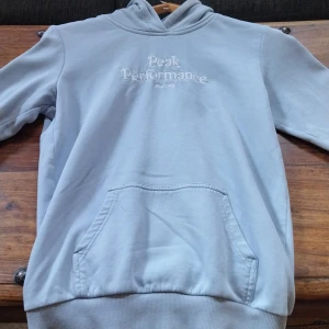 Ljusblå hoodie Peak Performance - Snygg ljusblå hoodie från Peak Performance med broderad logga på bröstet. Klassisk känguruficka framtill, huva och ribbade muddar. Perfekt för chill dagar eller när du vill ha en clean och enkel stil.