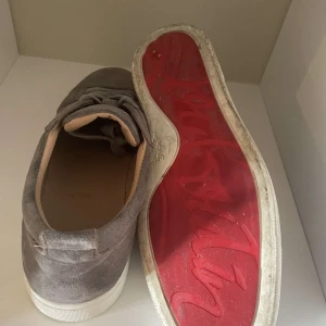 Grå mockasneakers från Louboutin - Snygga sneakers från Christian Louboutin i grå mocka med klassisk röd sula och vita kanter. Skorna har rund tå, snörning och en stilren design som passar till många outfits. Lyxig känsla med mjukt innerfoder och exklusiva detaljer.