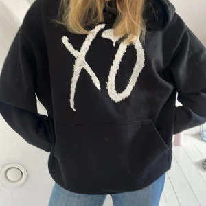 Svart The weeknd XO hoodie med stor logga - Svart hoodie från XO( The weeknds merch från hans konsert i Stockholm) med stor vit logga framtill. Tröjan har huva, känguruficka och en relaxed passform. Perfekt för dig som gillar streetwear och vill ha en skön och snygg tröja till jeans eller joggers.