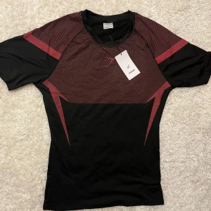 Svart och röd Gymshark t-shirt Onyx 5.0 - Sportig helt ny svart t-shirt från Gymshark med röda grafiska detaljer och mönster över bröst och rygg. Tillverkad i ett lätt och stretchigt syntetmaterial som andas, perfekt för träning. Kortärmad modell med rund hals och modern passform.