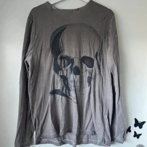 Grå långärmad tröja med dödskalle - Grå långärmad tröja från RG512 Rough Soul Co. med stor svart dödskalle-tryck på framsidan. Tröjan har en avslappnad passform och är tillverkad i mjuk bomull, perfekt för dig som gillar edgy streetstyle.