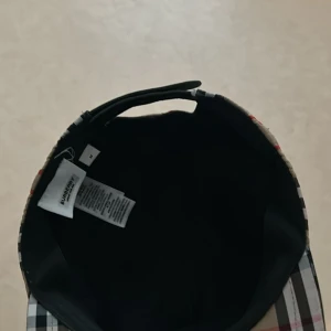 Burberry rutig keps med logga - Snygg keps från Burberry i klassiskt rutigt mönster med beige, svart, vitt och röda detaljer. Justerbar snapback baktill och tydlig Burberry London England-logga. Tillverkad i polyester, perfekt för dig som vill sticka ut med en ikonisk accessoar. Möts i goteborg bara.