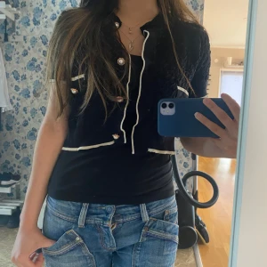 Svart elegant bolero med pärlknappar - Säljer en svart bolero med korta ärmar och vita kontrastkanter. Boleron har dekorativa pärlknappar framtill och två små fickor. Croppad modell som ger en trendig look, perfekt att styla över en t-shirt eller linne.