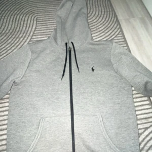 Grå hoodie från Ralph Lauren - Snygg grå hoodie från Ralph Lauren med dragkedja och svart logga på bröstet. Tröjan har huva med snörning och två stora fickor framtill. Perfekt för chill dagar och enkel att matcha med jeans eller joggers.