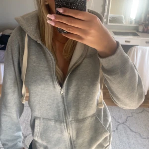 Grå russemerch dupe - Mysig grå hoodie med dragkedja framtill och stora fickor. Tröjan har huva med vita snören och är långärmad. Perfekt för chill dagar eller när du vill vara extra bekväm. Enkel och stilren design som passar till det mesta. Lite smutsig som inye går bort med bara tvättmaskin 