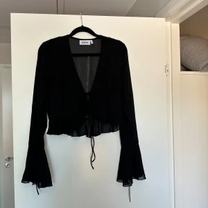 Svart blus med utsvängda ärmar Weekday - Säljer en svart, transparent blus från Weekday i storlek S. Blusen har v-ringning, knytning framtill och långa utsvängda ärmar med volangdetalj. Tunn och flowy känsla, perfekt för att lyfta vilken outfit som helst.