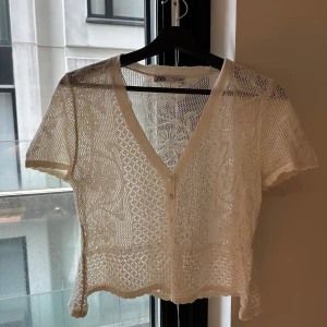 Vit spetsblus från Zara - Superfin vit blus från Zara med korta ärmar och v-ringning. Blusen är genombruten med spetsmönster och har knappar framtill. Perfekt för en somrig look med sina blommiga detaljer och lätta material.