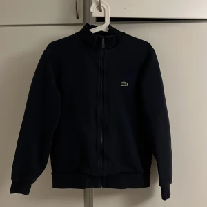 Svart zip från Lacoste - Säljer en svart zip från Lacoste med klassisk krokodillogga på bröstet. Tröjan har hög krage, hel dragkedja och ribbade muddar. Perfekt för dig som gillar stilren och sportig look. Materialet känns mjukt och bekvämt.