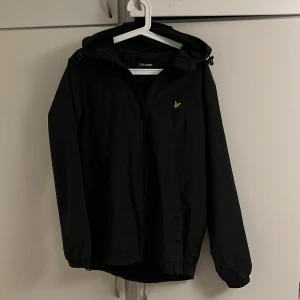 Svart vindjacka från Lyle & Scott - Svart vindjacka från Lyle & Scott med huva och dragkedja framtill. Jackan har en liten gul logga på bröstet och justerbar nederkant. Perfekt för blåsiga dagar och enkel att matcha med det mesta.