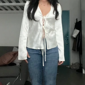 Slutsåld vit topp i satin med snörning - Säljer en superfin vit omlottopp i glansigt satinliknande material. Toppen har långa ärmar, djup v-ringning och snygg snörning framtill som ger en trendig look. Perfekt att styla med jeans eller kjol för en chic vibe.