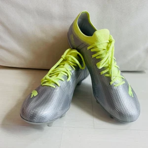 Nya Puma Ultra fotbollsskor, strl 38 - Nya snygga Puma Ultra fotbollsskor i metallic silver med neongula detaljer och snörning. Skorna har låg profil, rund tå och platt sula med fasta dobbar. Oanvända, endast provade. Ordinarie pris 1399kr