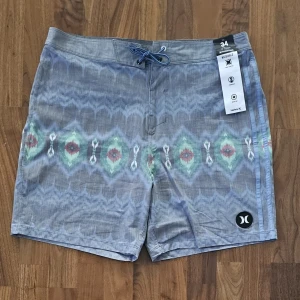 Blåmönstrade badshorts från Hurley - Snygga blå badshorts från Hurley Phantom med vågigt mönster i grönt, rött och vitt. Shortsen har snörning i midjan, bakficka med lock och är tillverkade i snabbtorkande, stretchigt återvunnet material. Perfekta för stranden eller poolen!