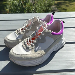 Vita chunky sneakers med lila detaljer - Snygga chunky sneakers i vitt med beige mockadetaljer och lila kant runt ankeln. Insidan är orange för en cool kontrast. Skorna har snörning och en tjock, platt sula som ger en sportig vibe. Perfekta för dig som gillar att sticka ut med färg och stil.