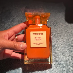 Tom Ford Bitter Peach parfym 100ml - Tom Ford Bitter Peach Eau de Parfum i en snygg, fyrkantig orange glasflaska på 100 ml. Flaskan har en exklusiv design med tydlig etikett framtill och en matchande orange kork. Doften är känd för sina fruktiga och sensuella toner. Bara testad 