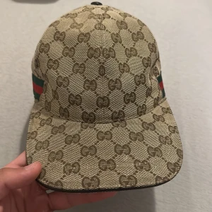 Gucci keps beige monogram S - Säljer en beige keps från Gucci med klassiskt GG-monogram över hela kepsen. Kepsen har gröna och röda ränder på sidorna och justerbart spänne bak. Tillverkad i Italien och i bomullsmaterial. Perfekt för dig som vill sticka ut med en lyxig accessoar.