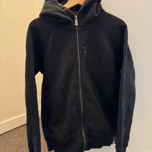 Svart hoodie från Sail Racing - Svart hoodie med dragkedja från Sail Racing. Tröjan har huva, lång ärm och en bröstficka med dragkedja. På ryggen finns en broderad logga. Perfekt för en avslappnad stil. Grejen till dragkedjan på fickan har lossnat